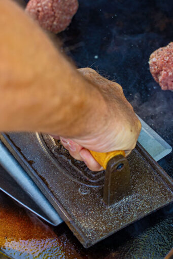 hands pressing down of a ground beef ball on a hot griddle with a spatula and weight to make a smash burger