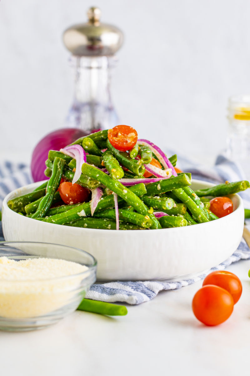 side view of a bowl of green bean salad in a white serving bowl next to a bowl of grated parmesan