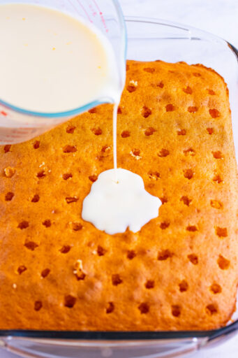 soak mixture being poured over slightly cooled orange cake with holes poked all over the top