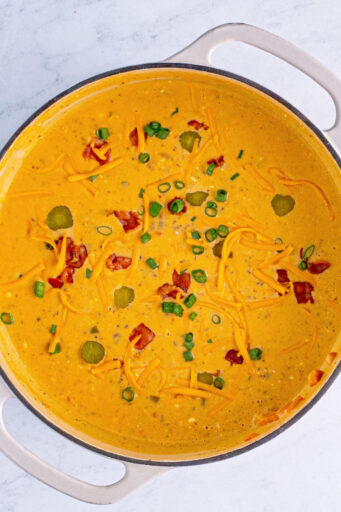 looking down at bacon cheeseburger soup in a pot topped with bacon, shredded cheddar, green onion, and pickles