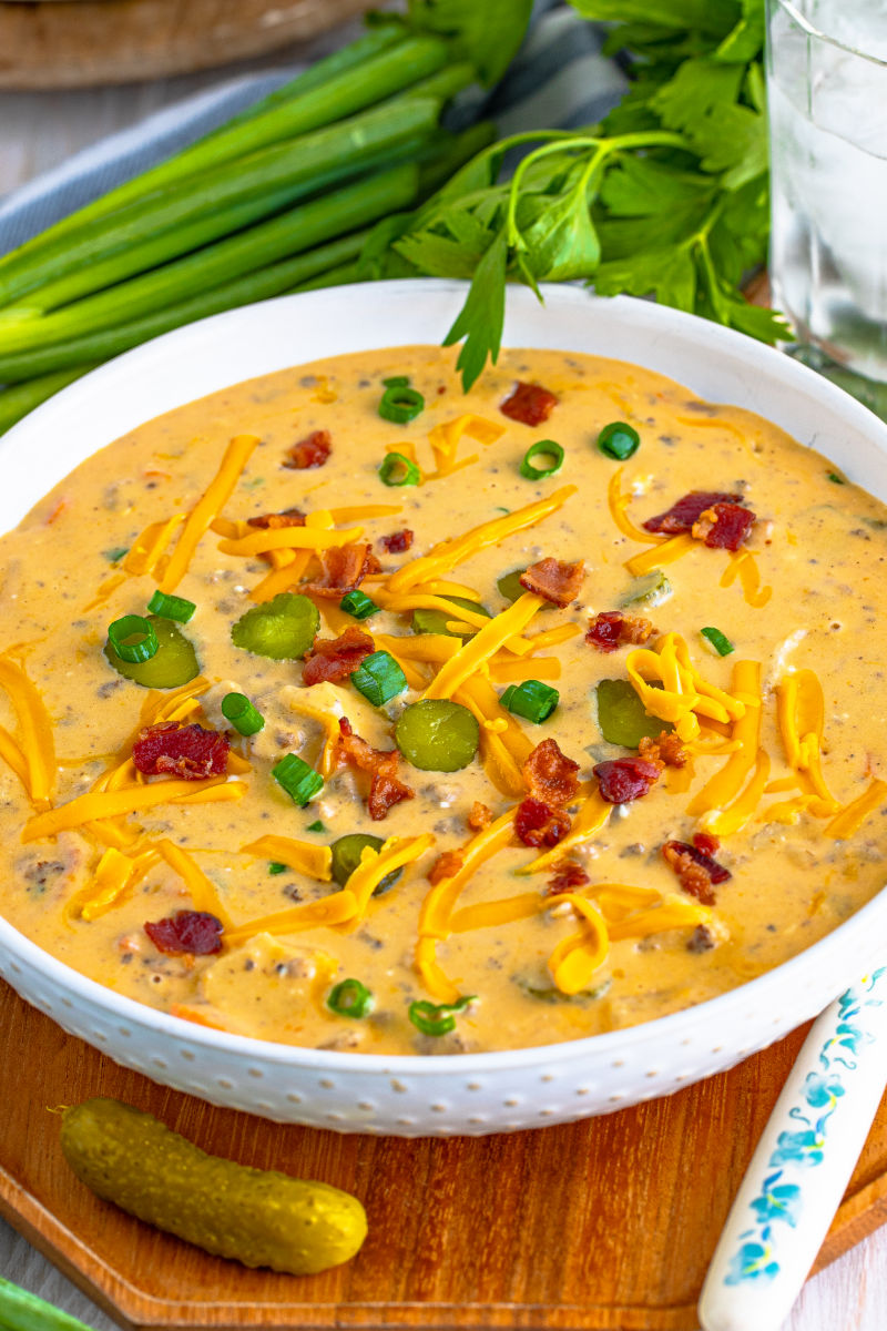 angled view of a bowl of bacon cheeseburger soup next to a spoon, a pickle, and green onions