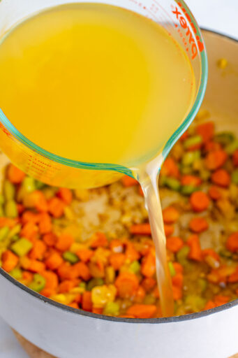 chicken broth being poured into a large pot with sautéed mirepoix