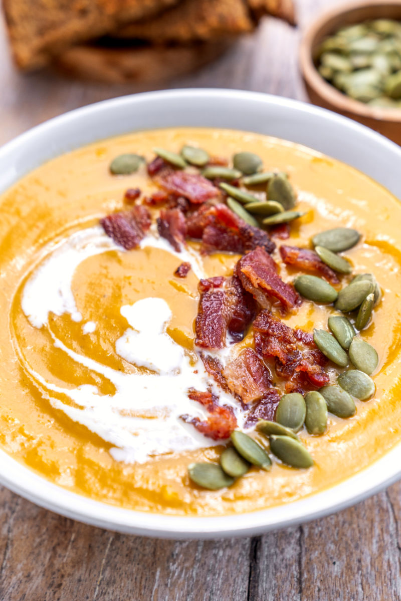 angled view of sweet potato soup in a bowl topped with cream, bacon, and pumpkin seeds